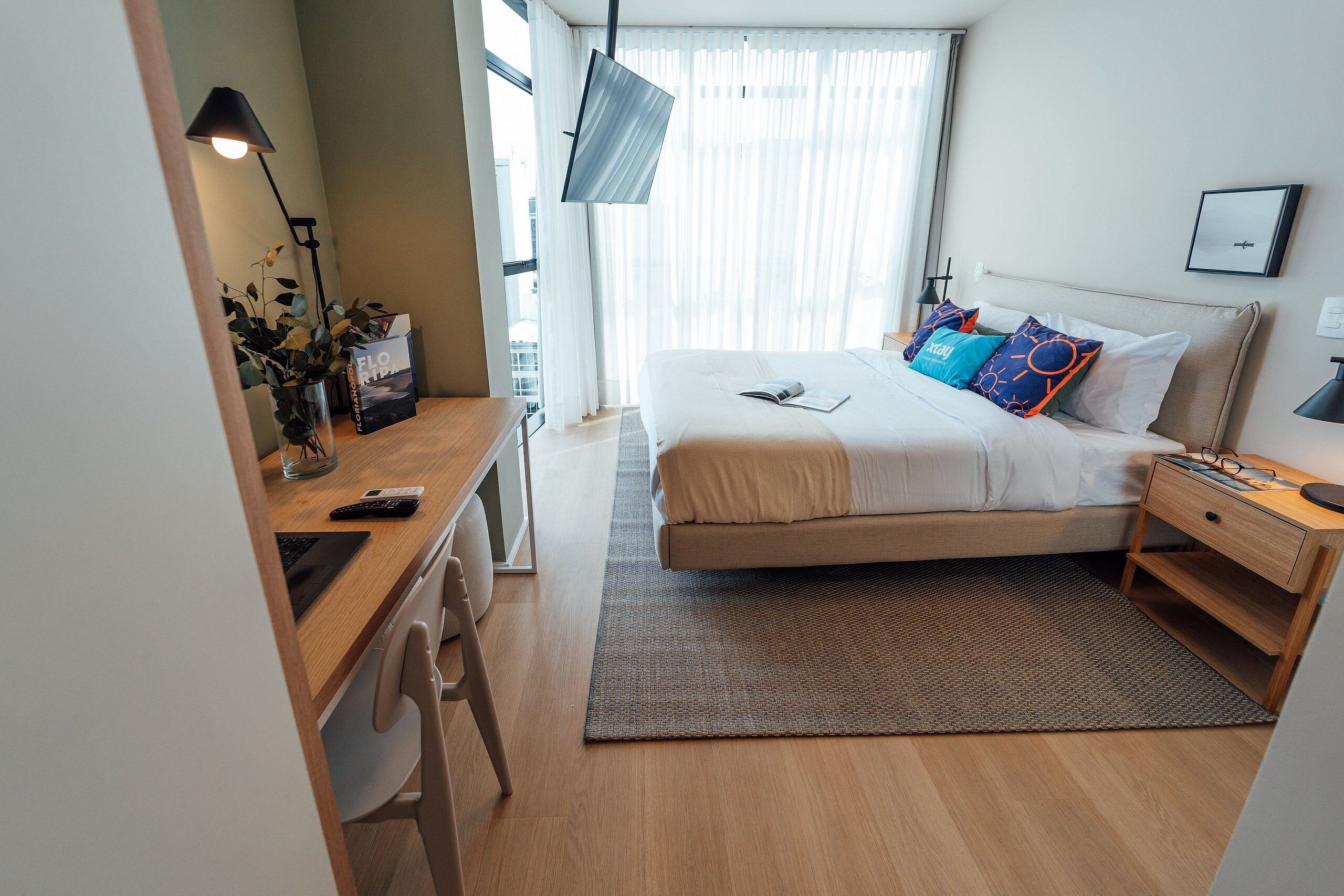 Ein modernes Hotelzimmer mit Bett, Nachttisch, Schreibtisch, Stuhl und einem großen Fenster mit Vorhängen.
