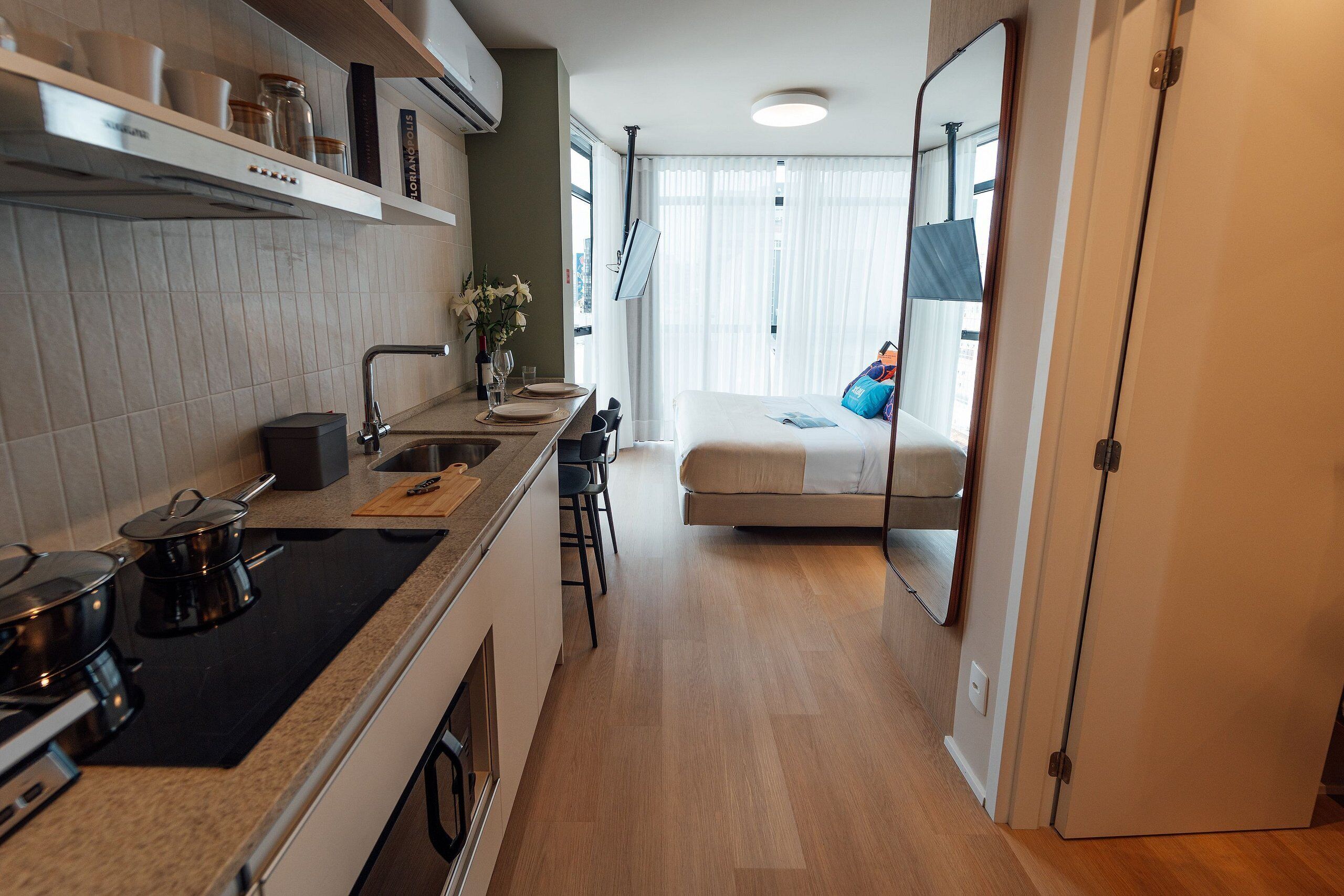 Cocina pequeña con estufa, lavamanos y taburetes de bar. Se ve una cama con almohada azul a través de una puerta abierta.
