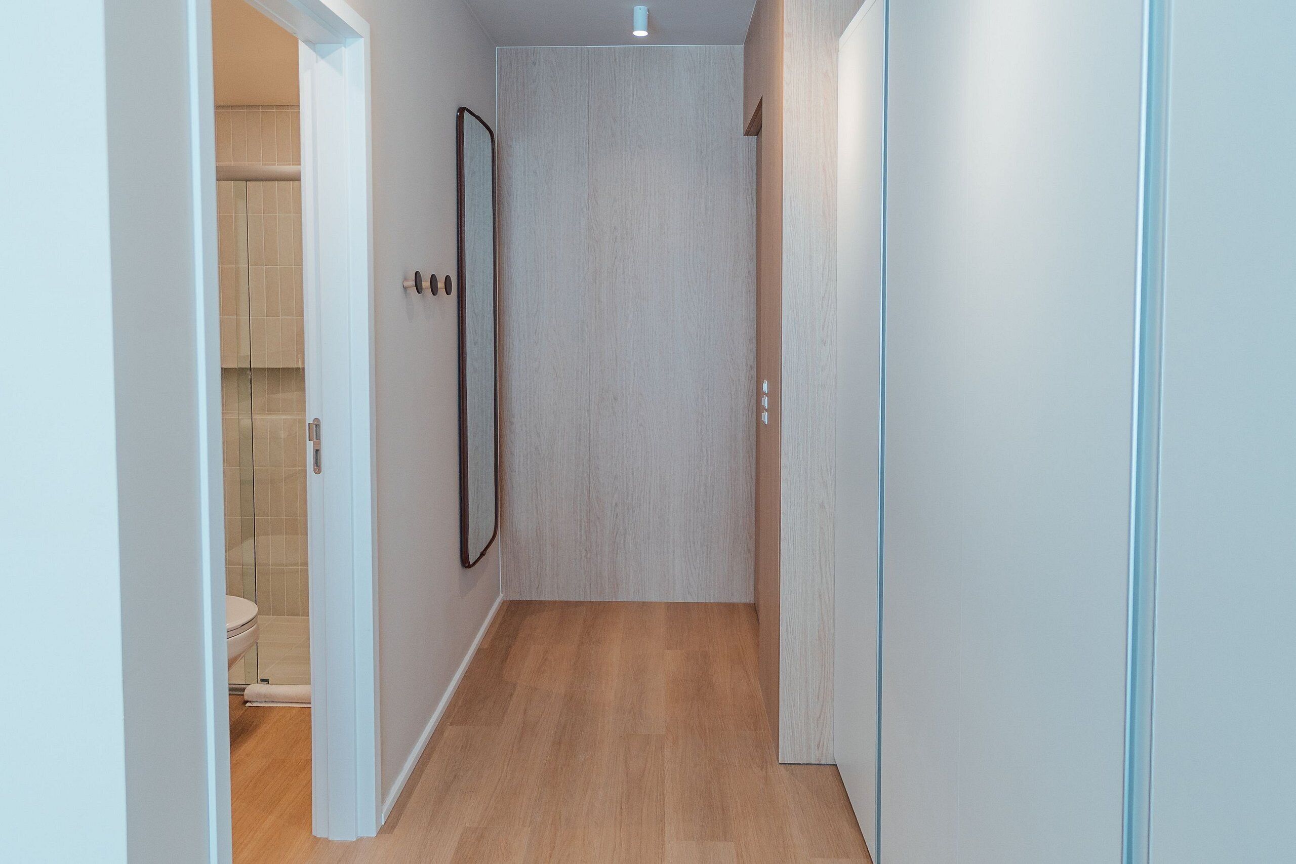 Un pasillo estrecho con suelo de madera, una puerta que da al baño y un espejo en la pared.