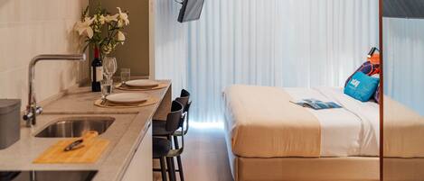 A modern hotel room with a kitchenette, a bed with a blue pillow, and a TV mounted on the wall.