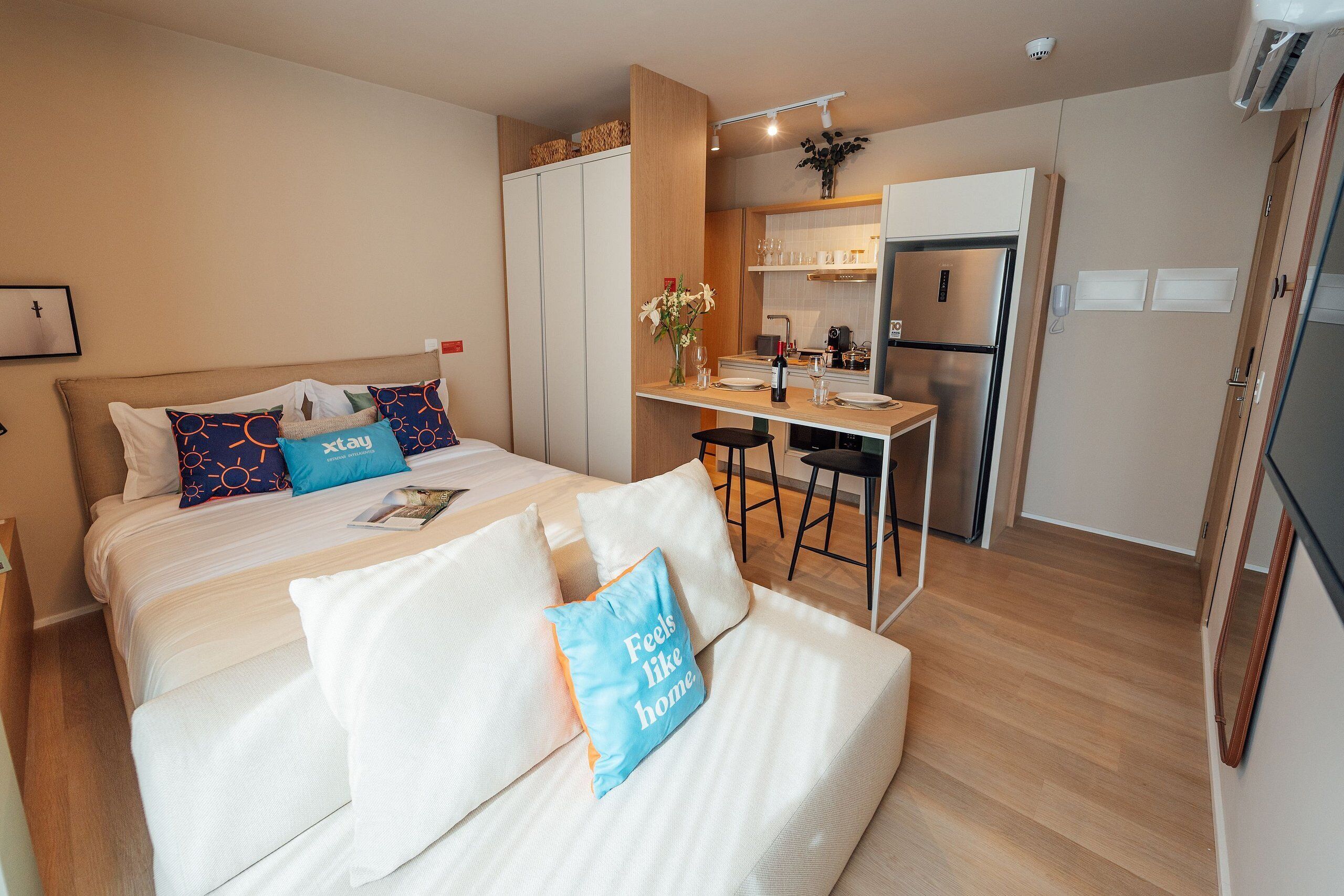 Quarto de hotel compacto com cama, pequeno espaço de cozinha e área de refeições.