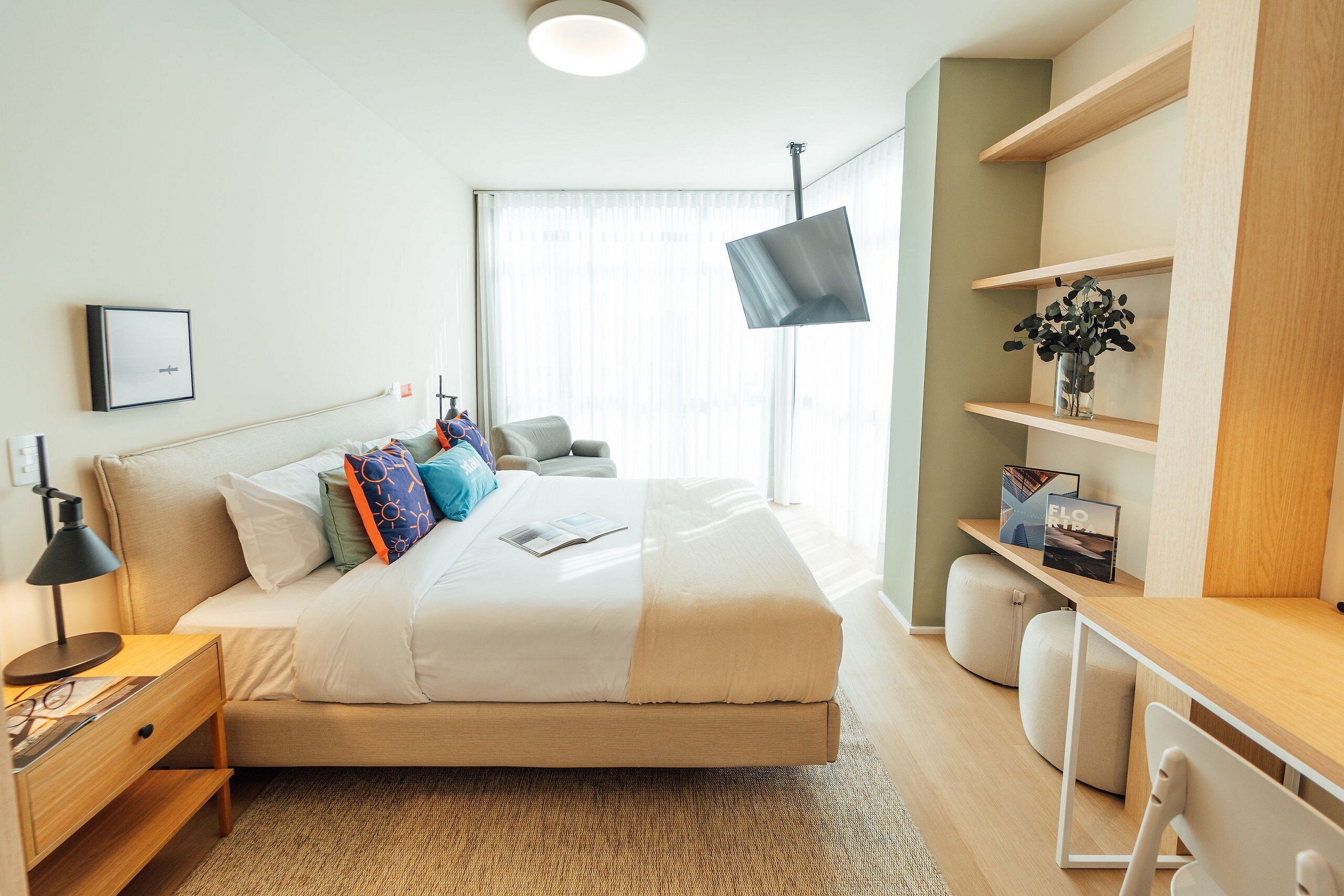 A modern hotel room with a bed, bedside table, desk, and shelves.