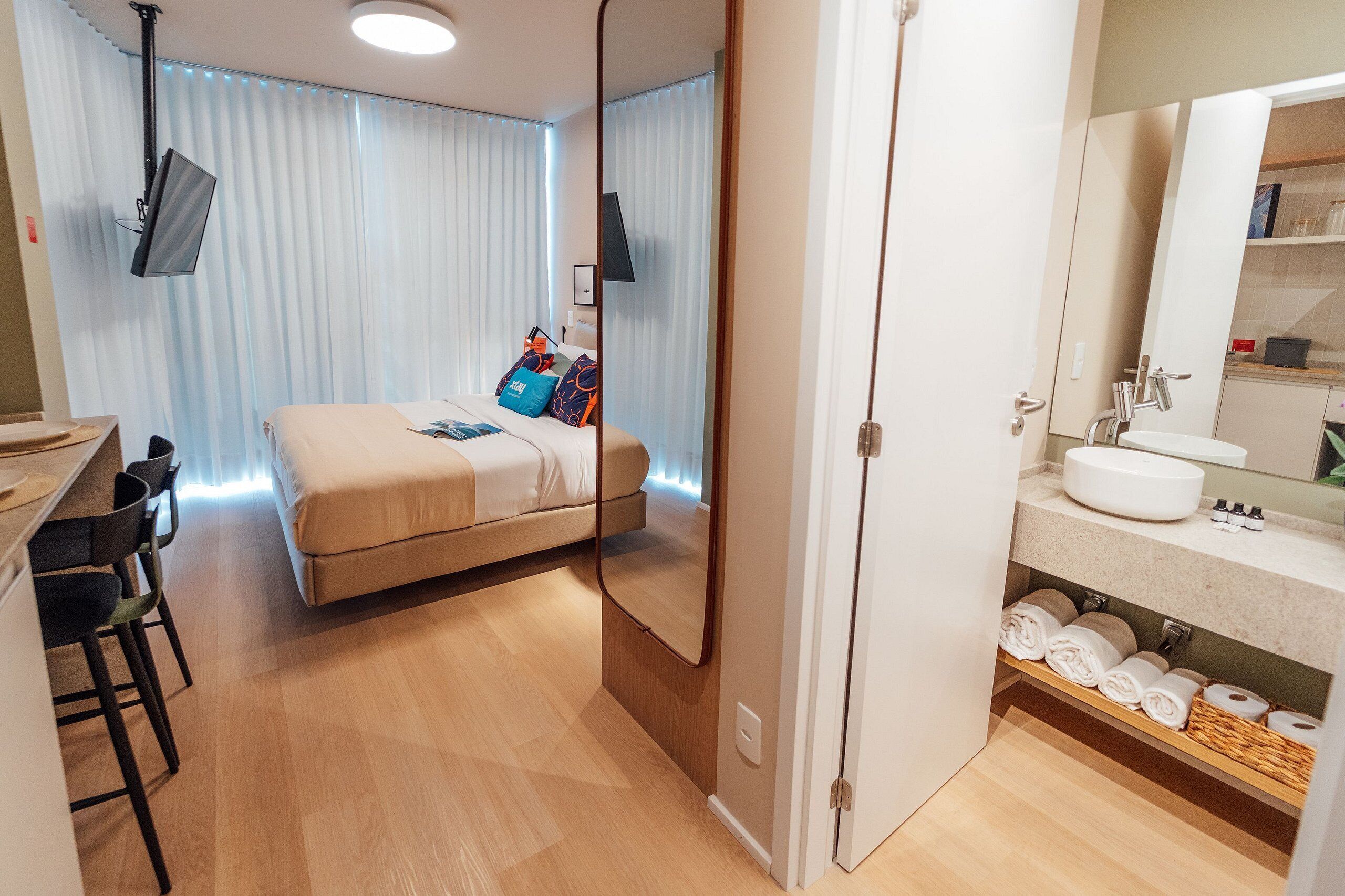 Une chambre d’hôtel comprenant un lit, un bureau avec des chaises, une salle de bain avec un lavabo et un miroir, et un téléviseur fixé au mur.