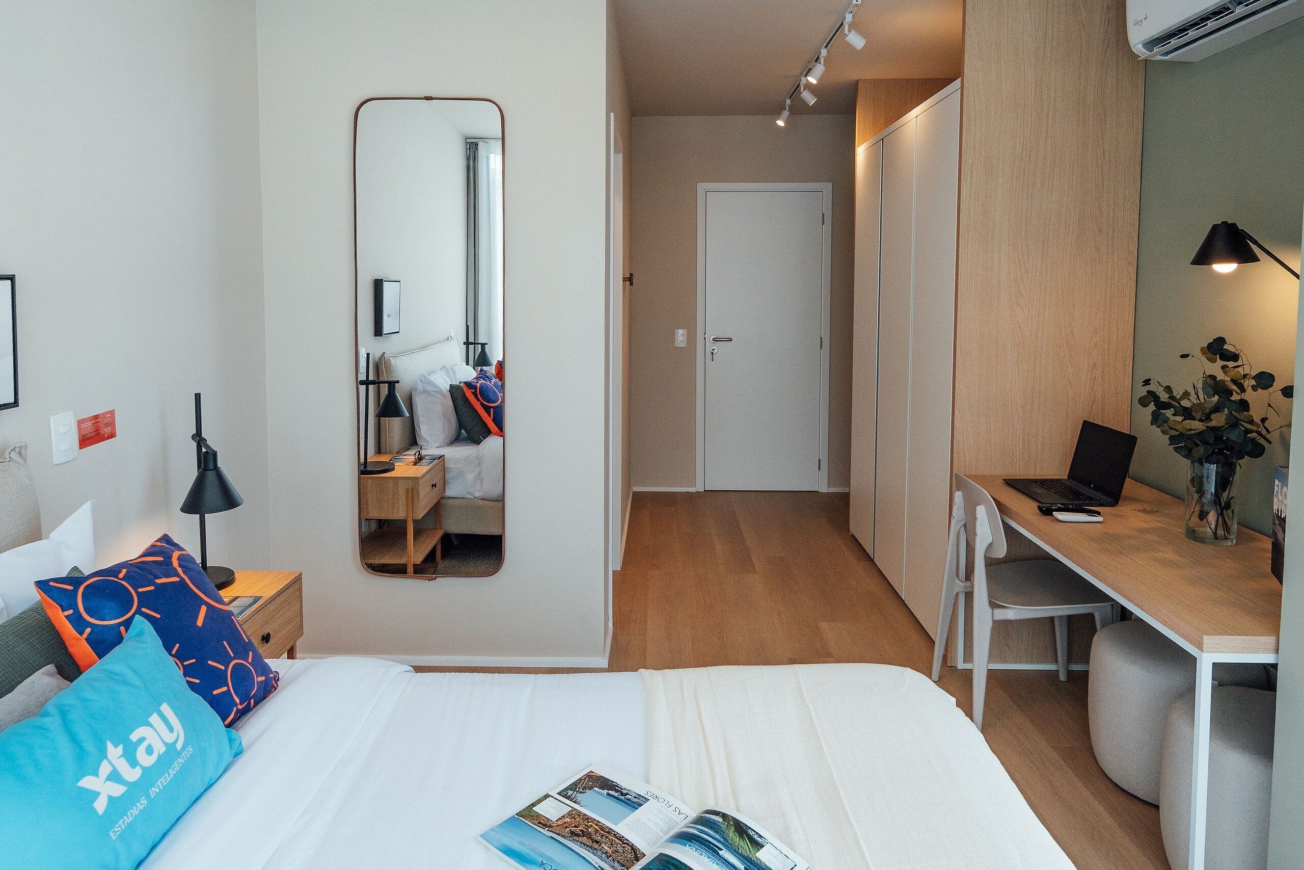 Une chambre d’hôtel moderne équipée d’un lit, d’une table de chevet, d’une lampe, d’un miroir, d’une armoire, d’un bureau, d’une chaise et d’un ordinateur portable.