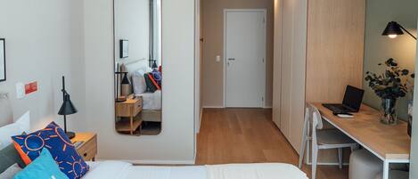 A modern hotel room with a bed, bedside table, lamp, mirror, wardrobe, desk, chair, and a laptop.