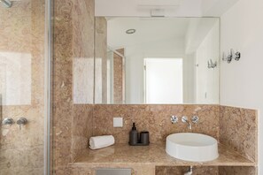 A modern bathroom with a marble countertop, a white sink, a large mirror, and a shower area. - Bridgeview Alcântara – The 3BR Loft Edition (Lisbon)
