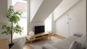 A modern living room with a flat-screen TV on a wooden stand, a grey sofa, a white chair, and a potted plant. - Bridgeview Alcântara – The 3BR Loft Edition (Lisbon)