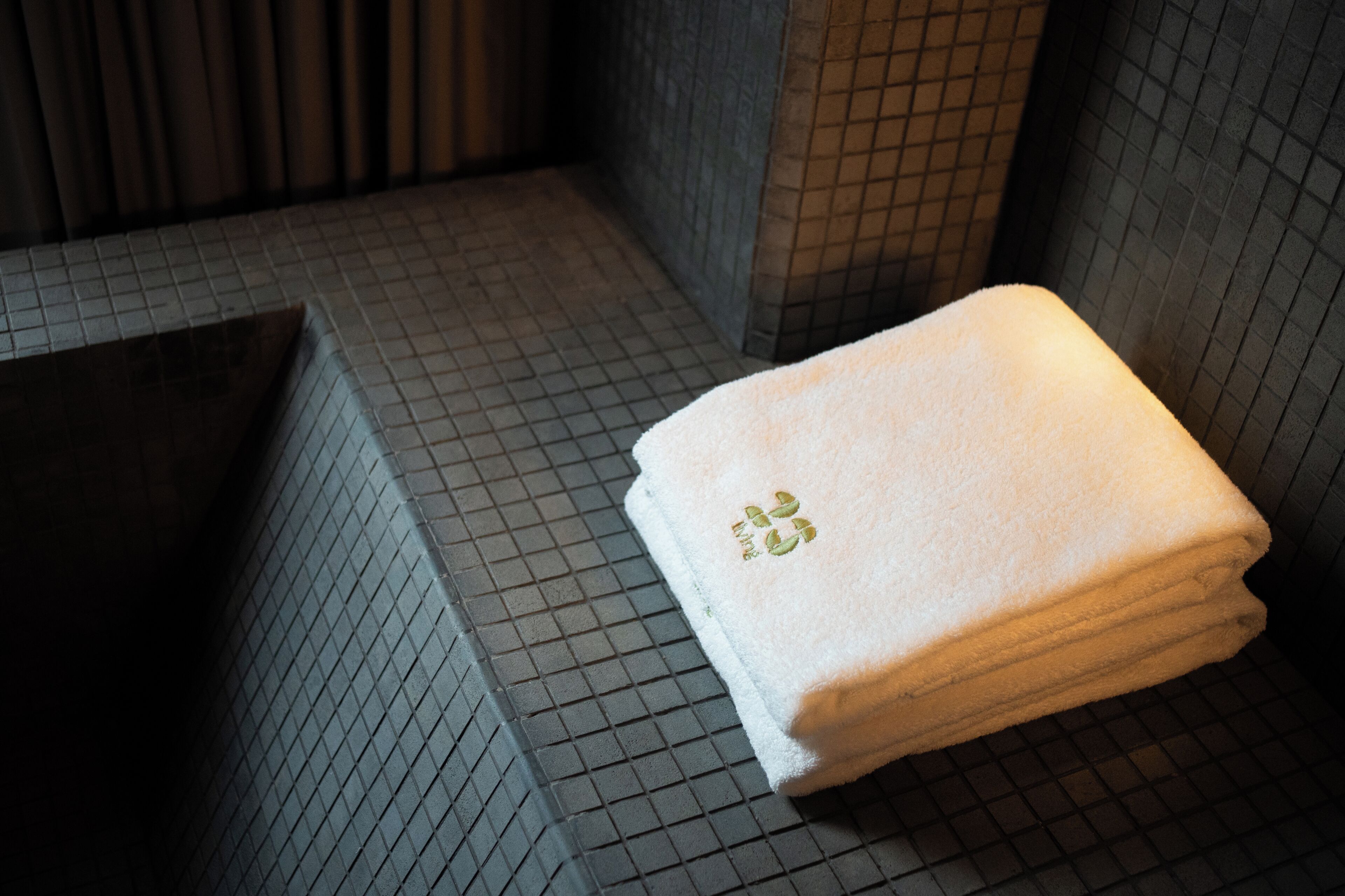 A bathroom with a tiled floor and walls, a towel with a logo, and a bench.