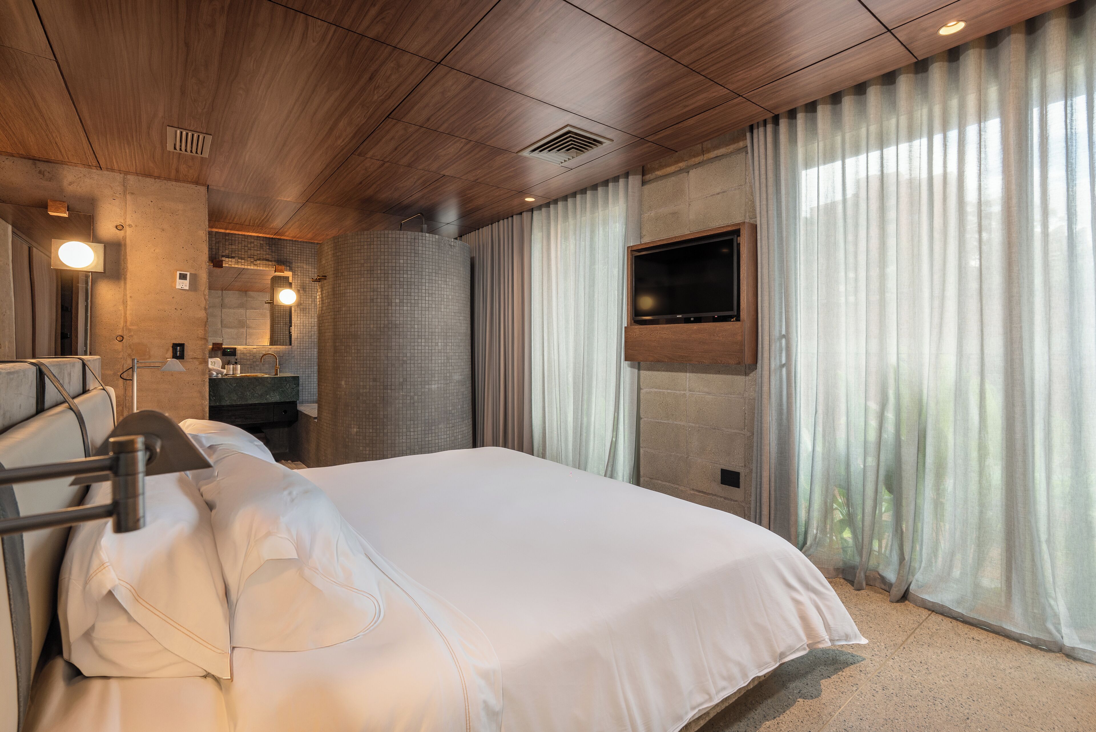 A hotel room with a wooden ceiling, a large bed with white bedding, a flat-screen TV mounted on the wall, a bathroom with a mirror and sink, and a window with sheer curtains.