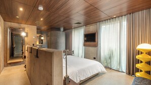 A modern hotel room with a wooden ceiling, a large bed, a TV mounted on the wall, and a bathroom with a large mirror and sink. - 23 Living (Medellín)