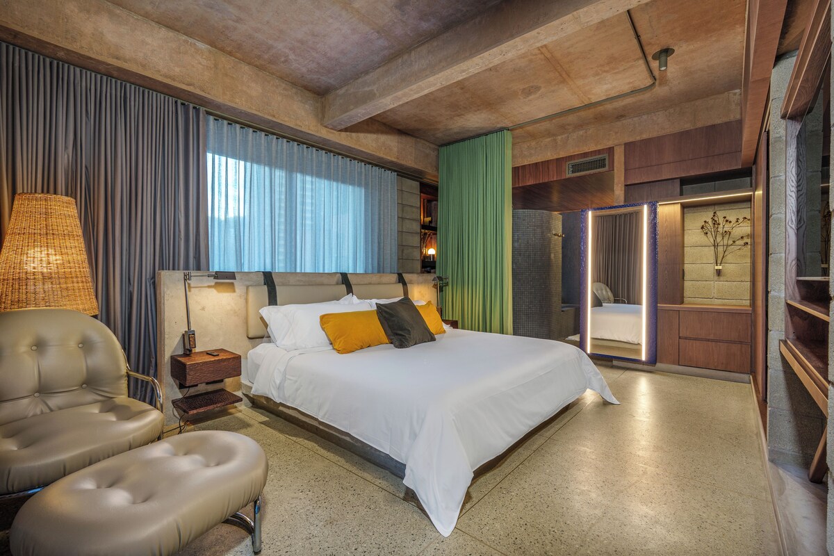 A modern hotel room with a large bed, a seating area with a chair and ottoman, a bedside table, and a view through a glass door.