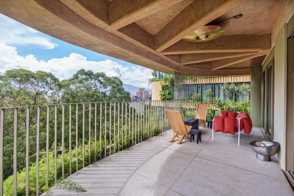 Uma varanda com vista para vegetação e edifícios, um cavalete de madeira, uma cadeira vermelha e uma tigela de metal.