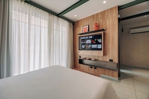 A hotel room with a large bed, a mounted TV, wooden paneling, and a window with sheer curtains. - GRES Apartamentos (Medellín)