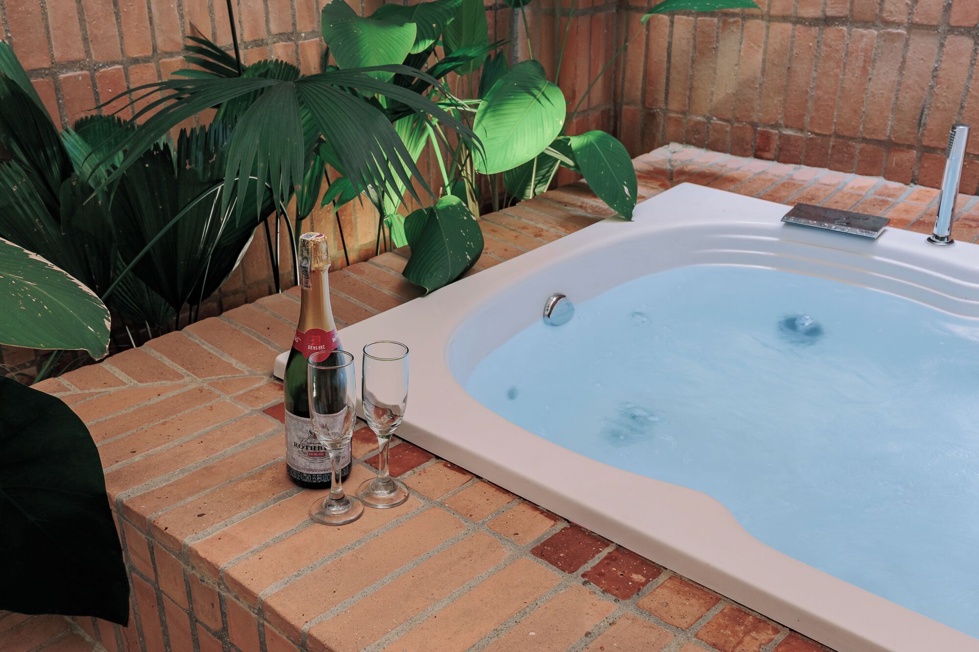 A hot tub with a brick border, a bottle of champagne, and two glasses on a table.