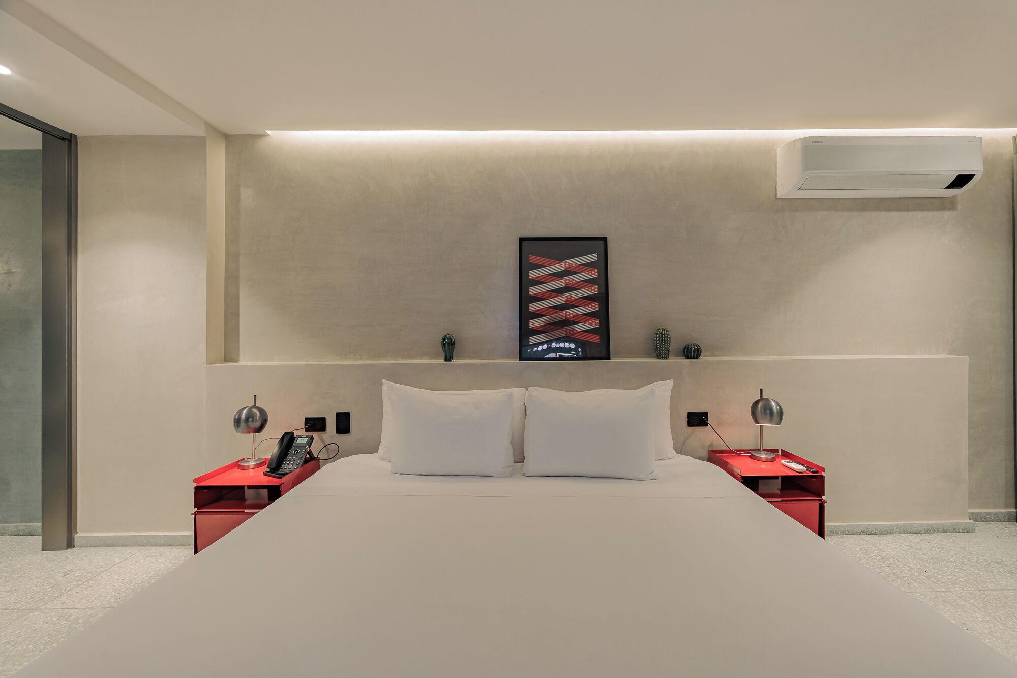 A modern hotel room with a large bed, two bedside tables with lamps, a mounted air conditioner, and a framed artwork on the wall.