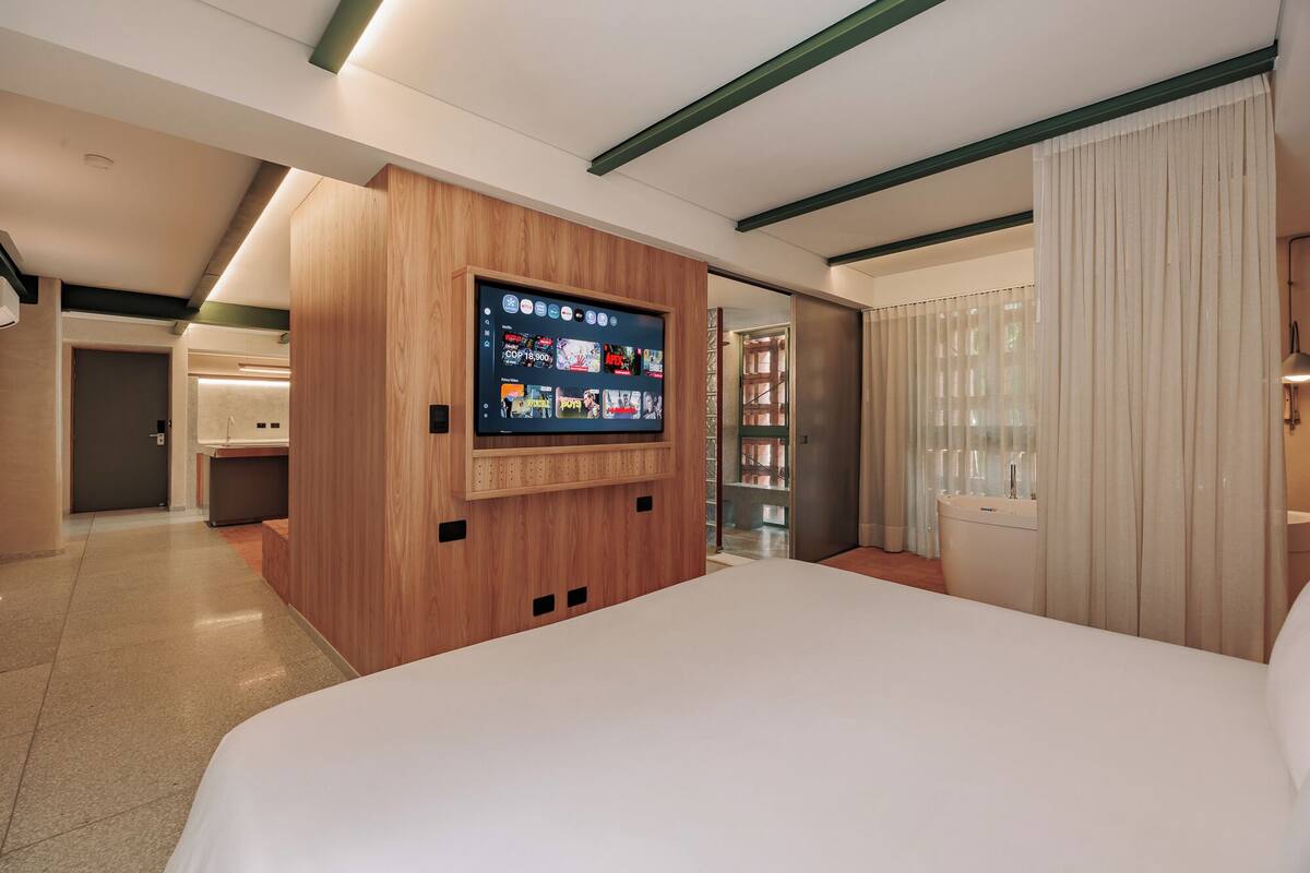 Una habitación de hotel moderna con una cama grande, un televisor montado en la pared, un baño con bañera y una zona separada con escritorio y silla.