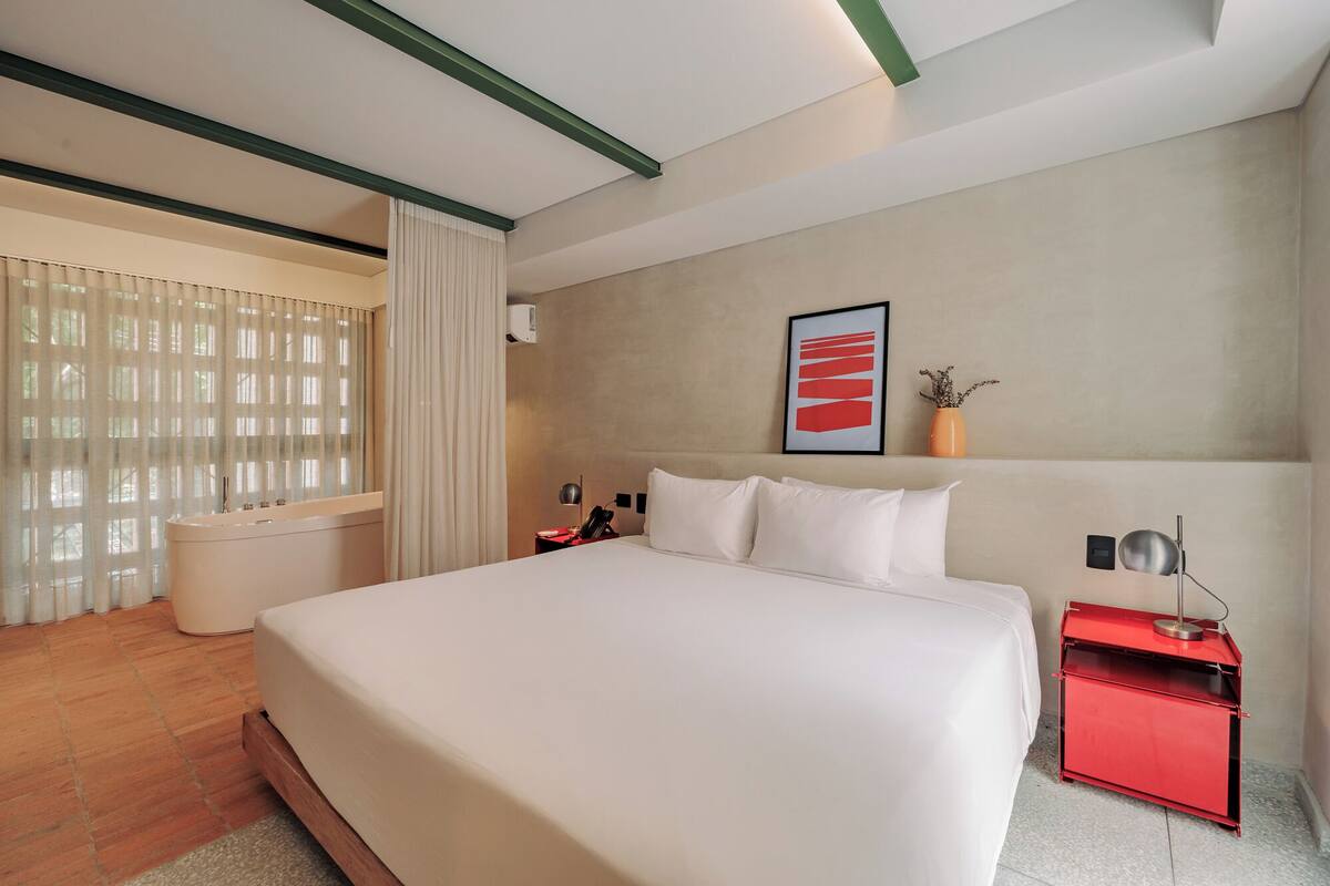 Habitación de hotel con una cama grande, una mesita de noche roja, una mesita de noche blanca, un aire acondicionado montado en la pared y una ventana con cortinas traslúcidas.