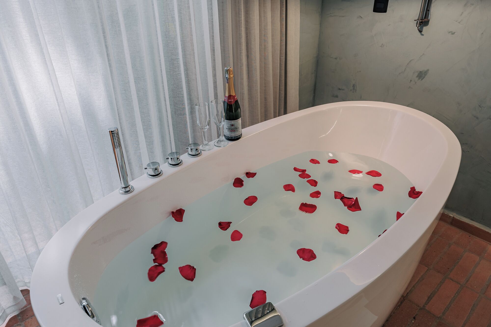 A bathtub with rose petals, a bottle of champagne, and a showerhead.