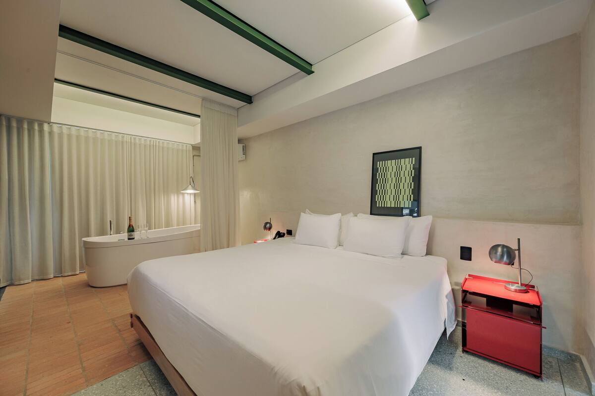 Habitación de hotel con una cama grande, una mesita de noche roja, un espejo y una obra de arte enmarcada en la pared.