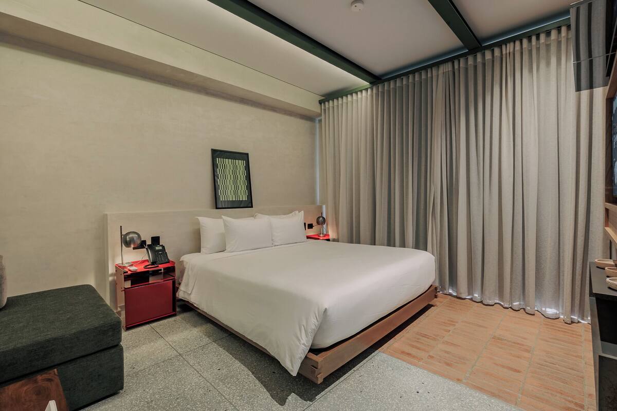 Una habitación de hotel moderna con una cama grande, una mesita de noche con lámpara, una pequeña mesita de noche roja, un sofá gris y un ventanal con cortinas.