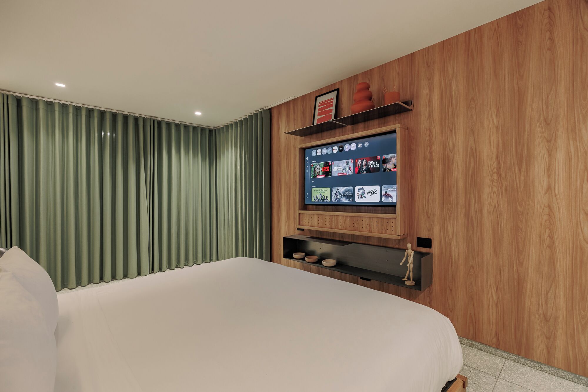 Quarto de hotel com uma cama grande, uma TV fixada exibindo vários canais e um painel de parede de madeira.