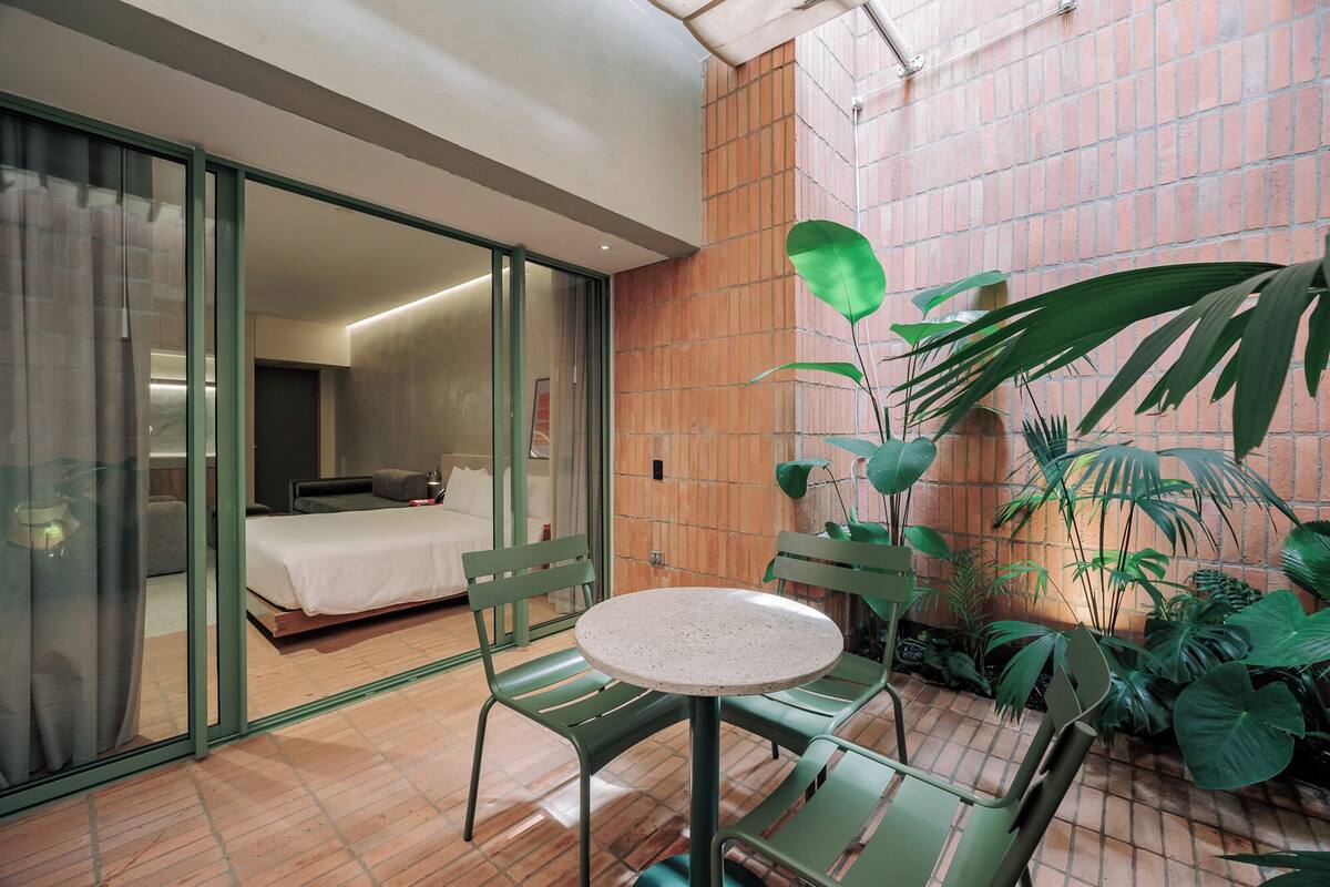 Habitación de hotel con cama, una silla verde, una mesita pequeña y un ventanal corredero que da a un balcón con plantas.