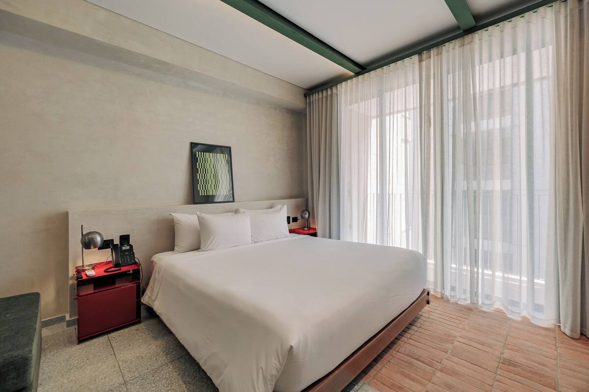 Habitación de hotel con una cama grande, un escritorio con lámpara, un cuadro en la pared y cortinas translúcidas en la ventana.