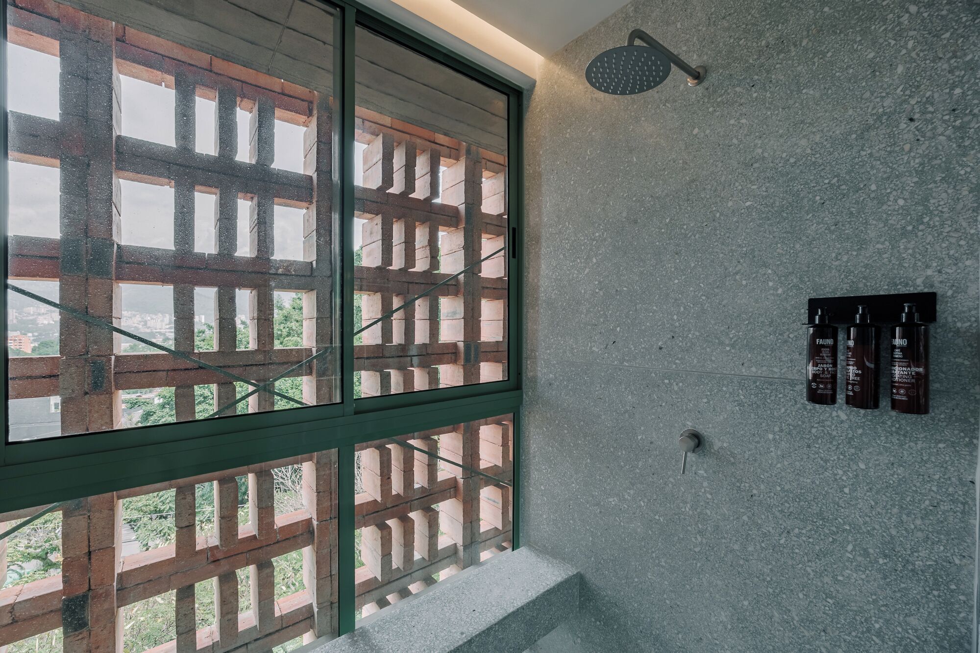 A bathroom with a shower, a window with a wooden lattice, and a wall-mounted soap dispenser.