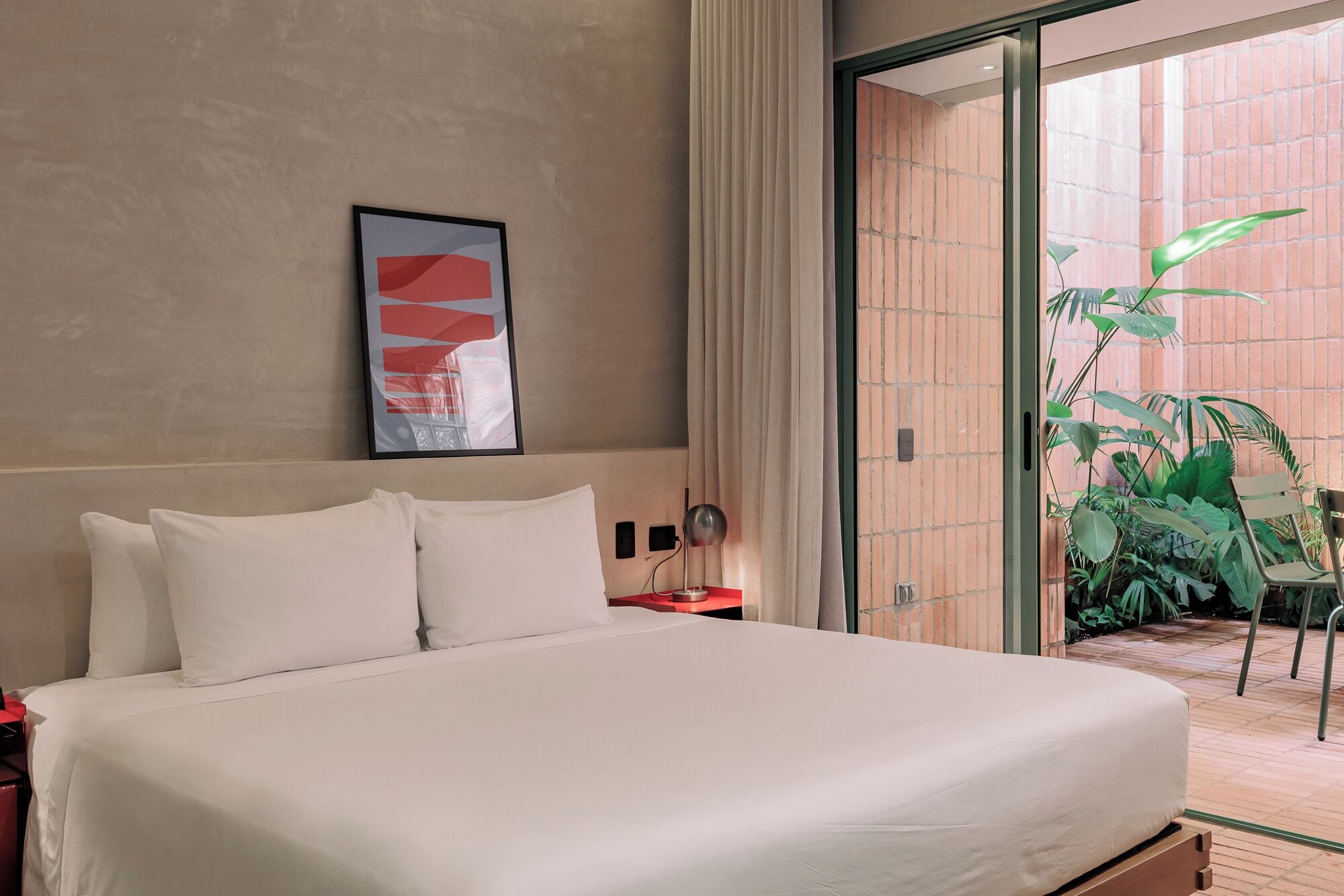 A hotel room with a bed, a nightstand, a lamp, a framed abstract artwork, and a view of a brick wall and greenery outside.