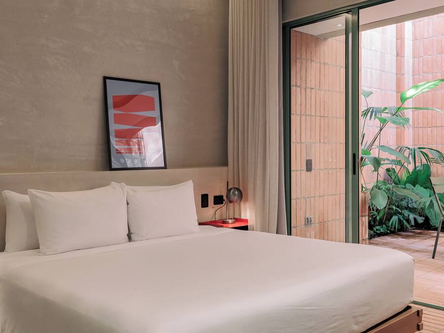 Ein Hotelzimmer mit Bett, Nachttisch, Lampe, einem gerahmten abstrakten Kunstwerk und Blick auf eine Ziegelwand und Grünpflanzen draußen.