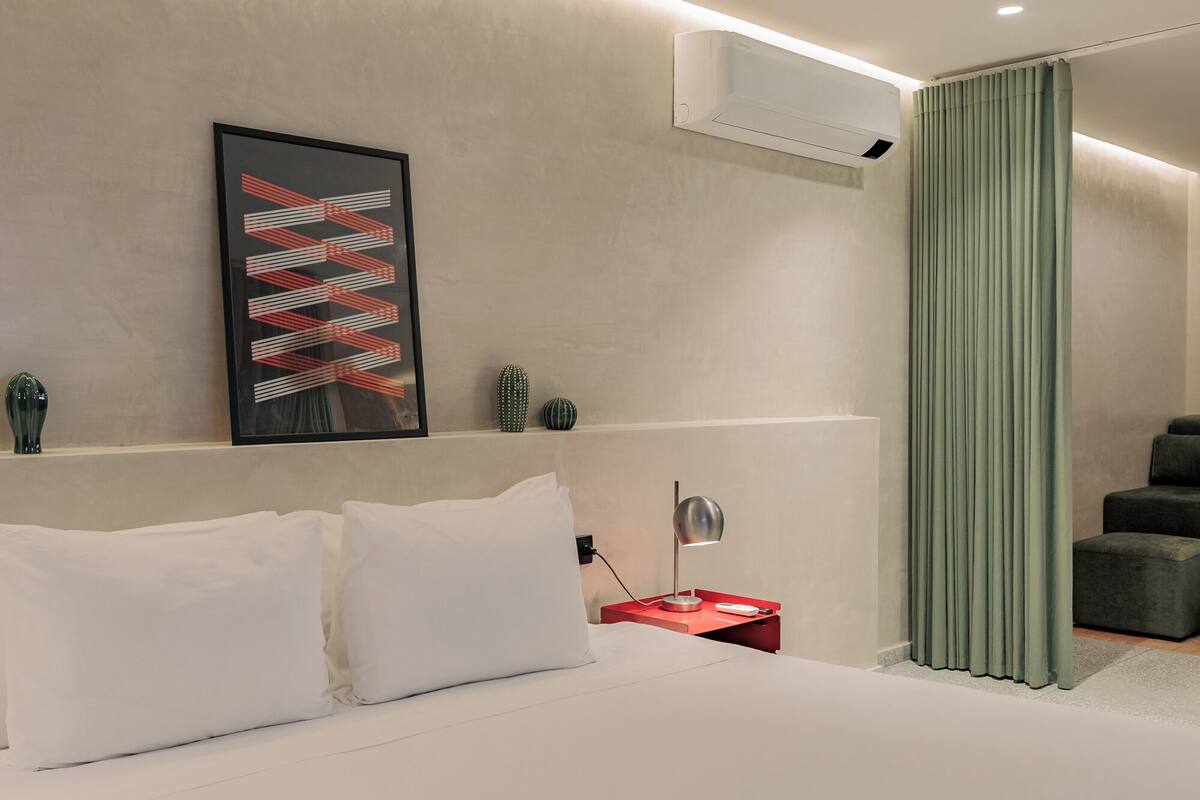 Una habitación de hotel moderna con una cama, una mesita de noche, una lámpara de escritorio, una obra de arte abstracta enmarcada, un equipo de aire acondicionado y una cortina verde.