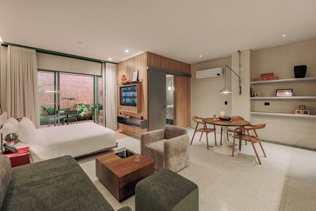 Una habitación de hotel moderna con cama, sofá, mesita de centro, televisor, zona de comedor con sillas y balcón con plantas.