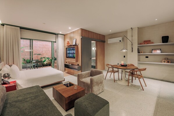 A modern hotel room with a bed, a sofa, a coffee table, a TV, a dining area with chairs, and a balcony with plants. - GRES Apartamentos (Medellín)