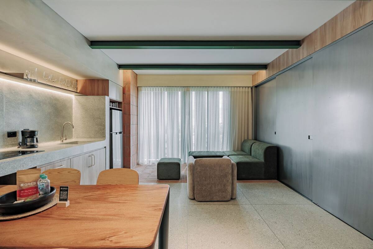 Habitación de hotel compacta con kitchenette, zona de comedor, espacio de estar con sofá y reposapiés, y un amplio ventanal corredero.