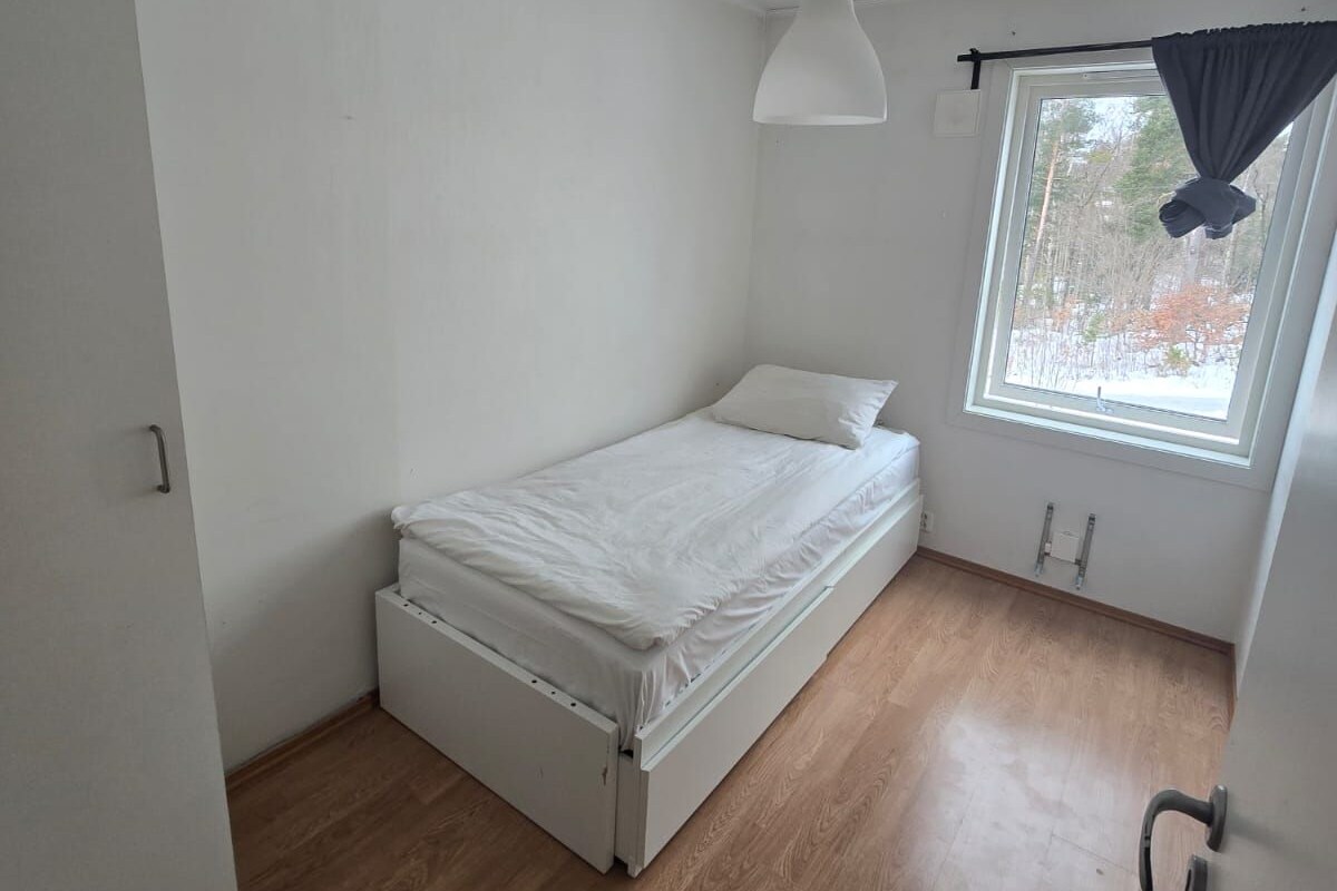 Ein kleines Zimmer mit einem Einzelbett, Holzboden, weißer Decke und einem Fenster mit Vorhang.