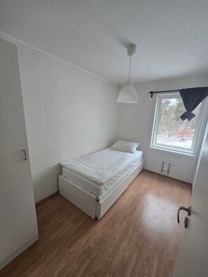 Ein kleines Zimmer mit einem Einzelbett, Holzboden, weißer Decke und einem Fenster mit Vorhang.