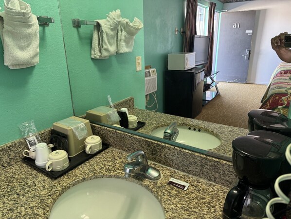 Baño de hotel con encimera de granito, lavamanos, espejo y cafetera.