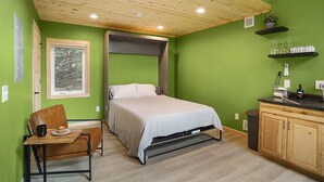 A bedroom with a bed, a wooden ceiling, a window with a view of trees, a small table with a mug and muffin, and a kitchenette with shelves and a trash can. - 2 Level Cabin w/Hot Tub & Fireplace. 1.5 Bathrooms, 2 Queen Beds (Ashland)