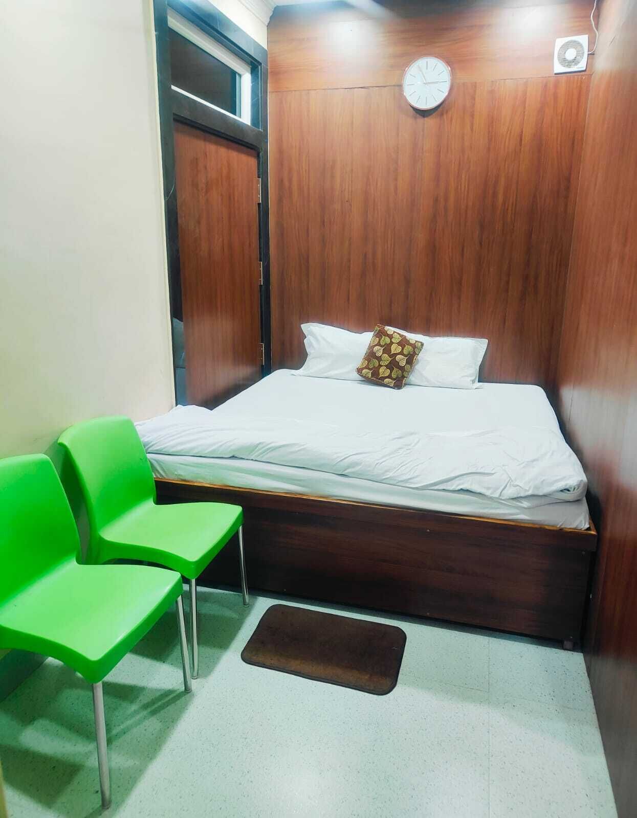 A small, neatly arranged room with a wooden headboard, a bed with white linens, two green chairs, and a small rug.