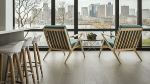 A modern room with large windows, two wooden chairs with cushions, a small table with a plant, and a row of bar stools.