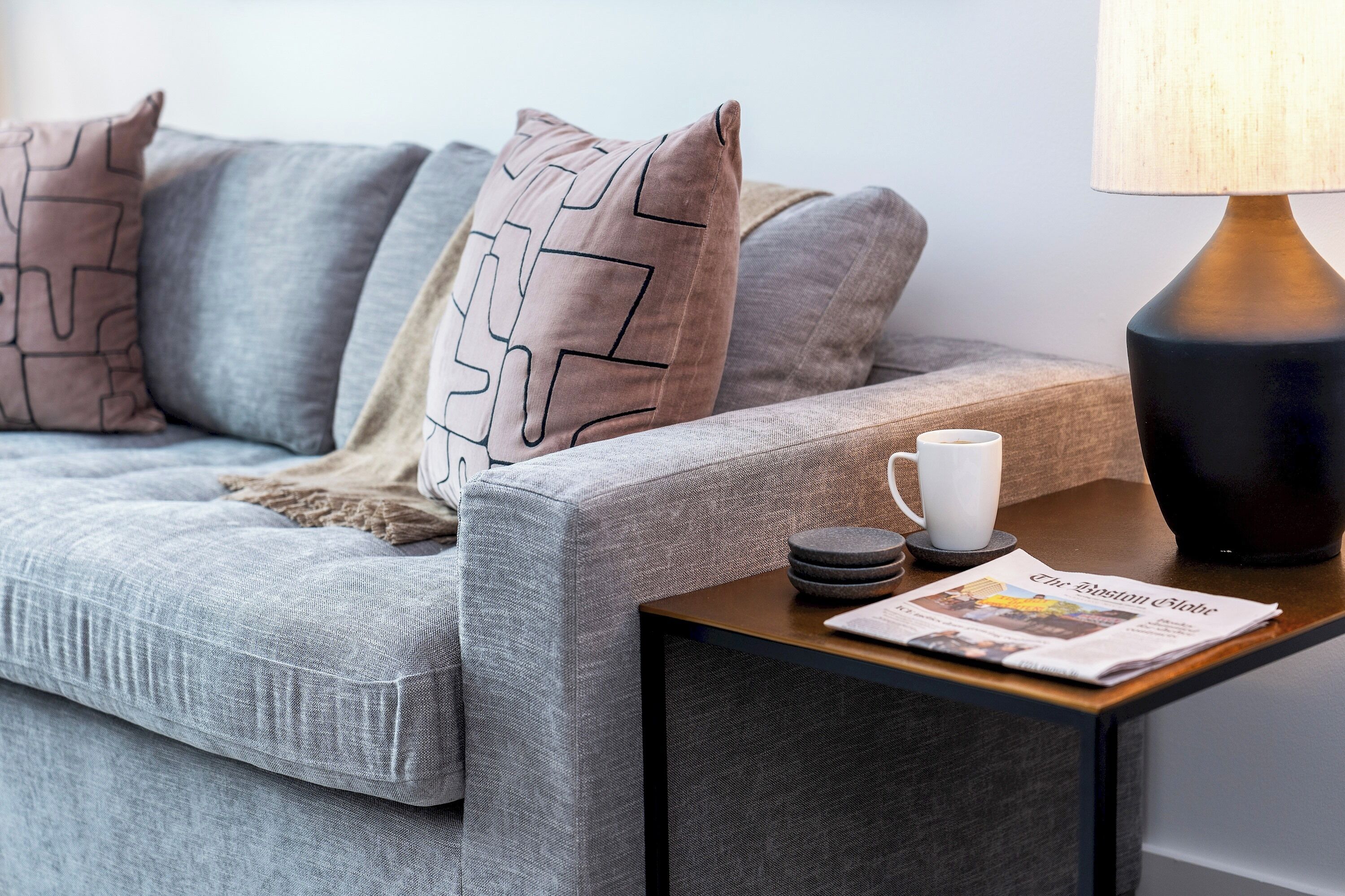 A modern living room with a grey sofa, patterned cushions, a coffee table with a newspaper, and a lamp.