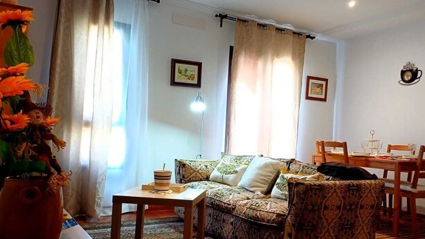 Un salon comprenant un canapé, une table basse et une table à manger avec des chaises. On y trouve des rideaux, une lampe et des tableaux encadrés accrochés aux murs.
