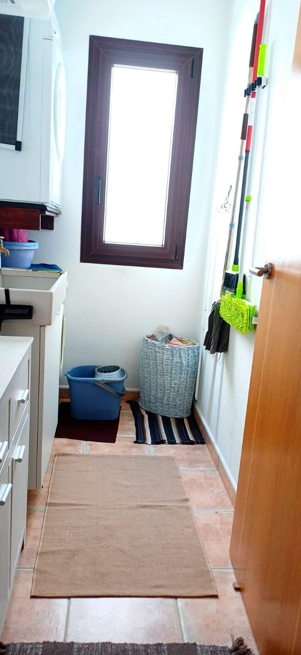 Una cocina estrecha con ventana, un cubo azul, una cesta de mimbre y una puerta de madera.