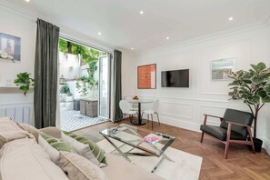 A modern living room with a glass coffee table, a sofa, a dining area with a table and chairs, a TV mounted on the wall, and a potted plant. - Lovely 1 bed Garden Flat - Earl s Court (London)