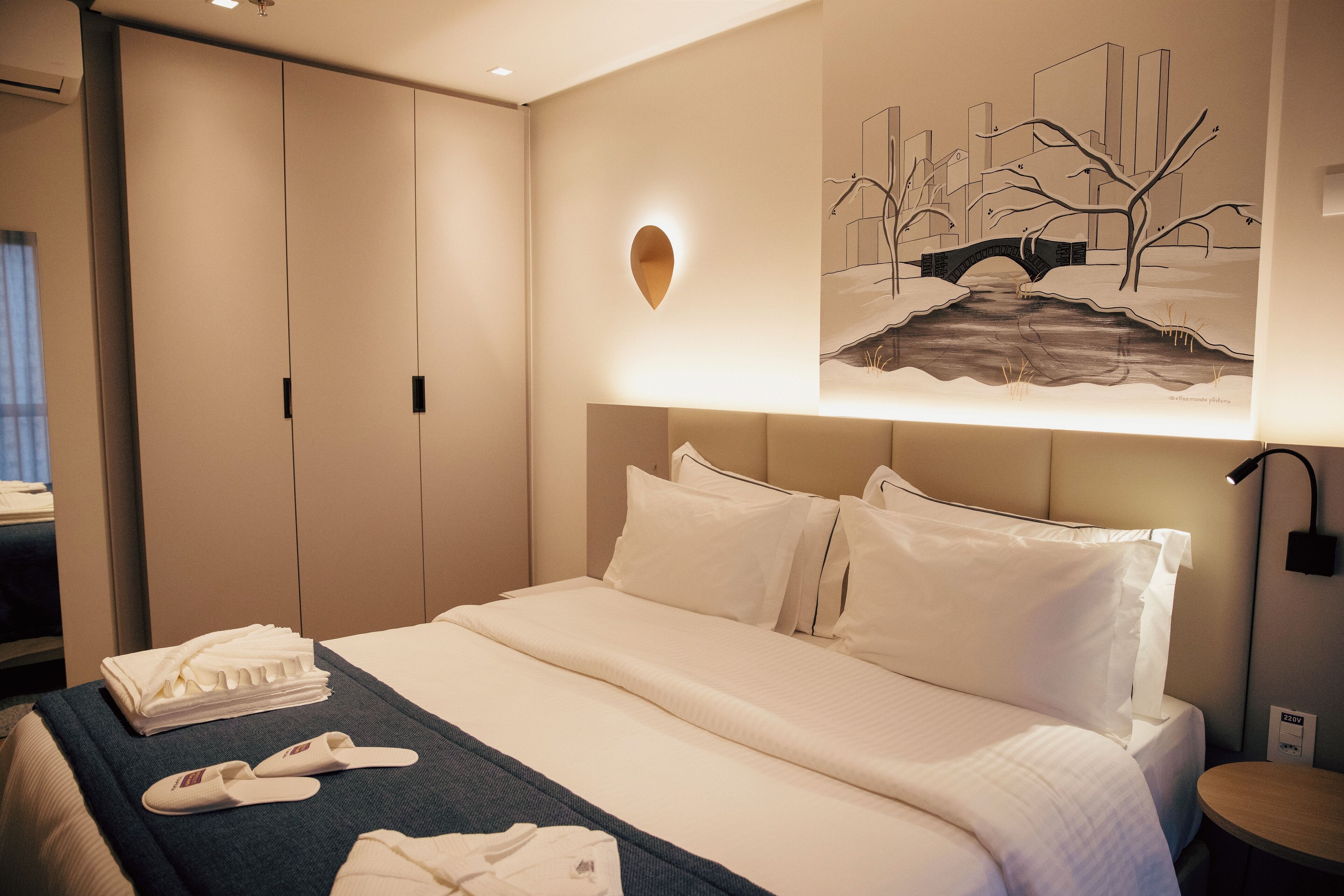 A hotel room with a neatly made bed, a bedside table, a wall-mounted light, and a painting of a bridge.