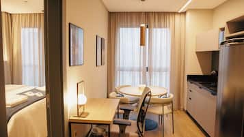 Una habitación de hotel con una cama, un escritorio con silla, una mesa de comedor con sillas, una cocina con refrigerador y una ventana con cortinas.