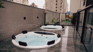 Two hot tubs in a modern outdoor setting with a glass enclosure, surrounded by a high-rise building and a play area in the background. - Nobile Grand Itapema (Itapema)