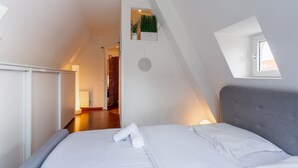A modern bedroom with a sloped ceiling, a bed with white linens, a grey upholstered headboard, a small window, and a wall-mounted plant. - Le Cocon Alsacien - Appt authentique coeur strasbourg (Strasbourg)