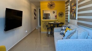 Una sala moderna con sofá, televisor de pantalla plana en la pared, comedor con mesa y sillas, y una cocina al fondo.
