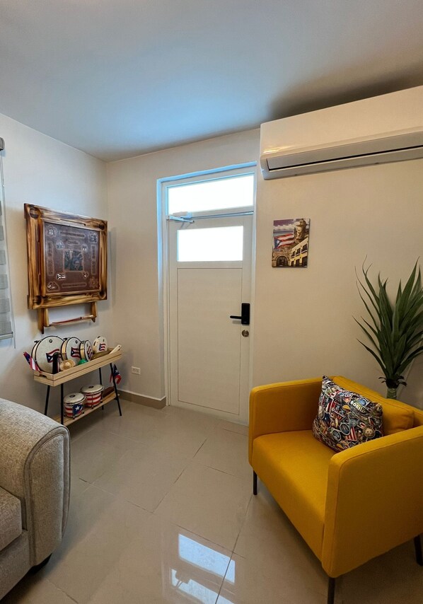 Una sala de estar con un sillón amarillo, una mesita con tambores, una puerta y una planta.